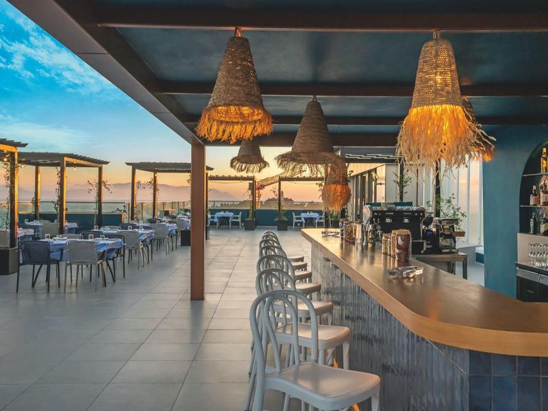Outdoor bar and dining area with stools, tables, and sunset view under a covered patio.