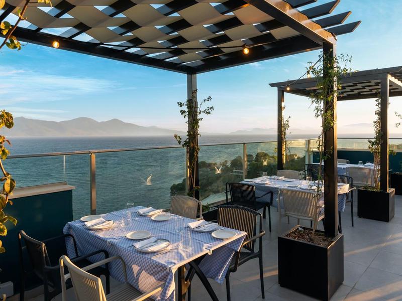 Outdoor dining area with tables and pergolas overlooking a scenic mountain and ocean view.