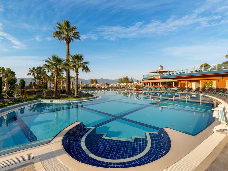 Resort pool with palm trees under a blue sky, surrounded by lounge chairs and a building.
