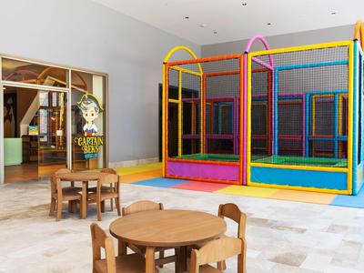Sala giochi luminosa per bambini con trampolino colorato e tavoli di legno con sedie.