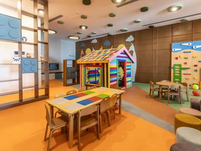 Area giochi colorata per bambini con tavoli, sedie e una casetta in una camera d'hotel.