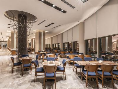 Ampia sala da pranzo con numerosi tavoli e sedie, illuminazione moderna e grandi finestre.