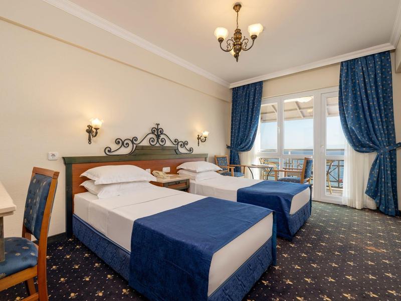 Hotelzimmer mit zwei Einzelbetten, blauen Bettdecken, Teppichboden und Balkon mit Meerblick.