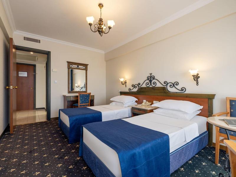 Hotelzimmer mit zwei Einzelbetten, blauen Decken, Teppichboden und klassischer Wandbeleuchtung.