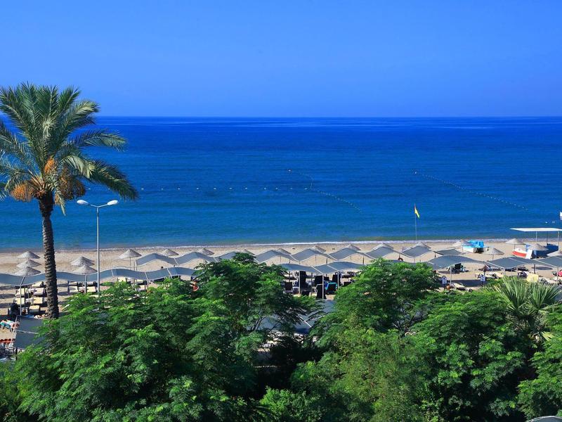 Strand mit Sonnenschirmen, blauer Himmel, Meer und grünen Palmen im Vordergrund.