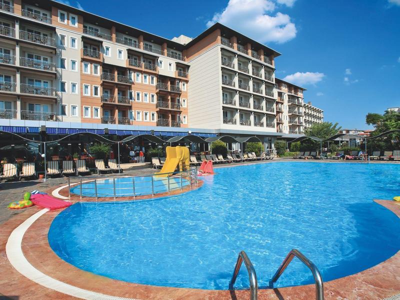 Großer Außenpool mit Rutsche vor mehrstöckigem Hotelgebäude bei sonnigem Wetter.