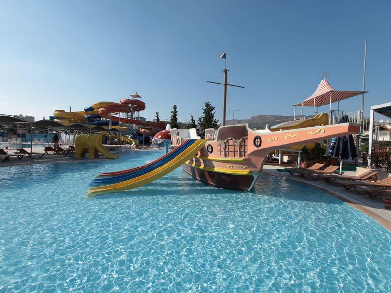Wasserpark mit großem Schwimmbecken, Rutschen und einem Spielschiff unter blauem Himmel.