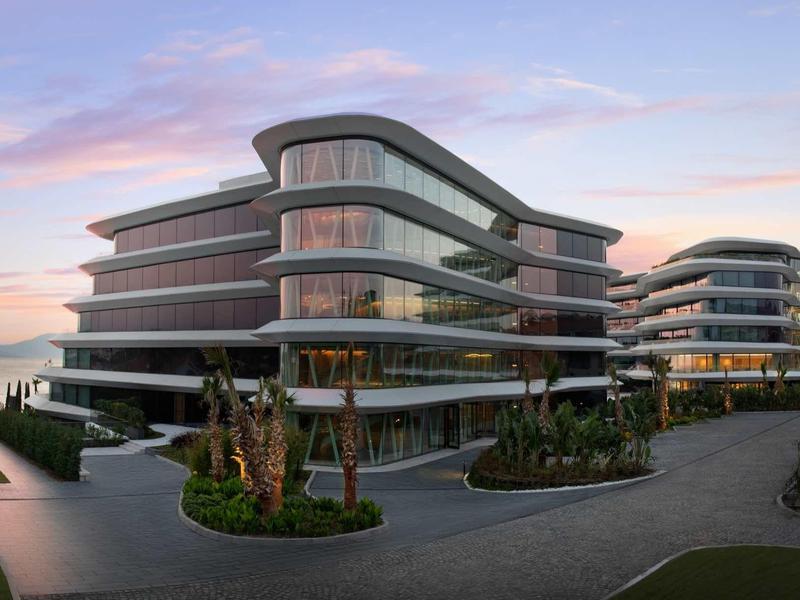 Moderne Bürogebäude mit abgerundeten Ecken, großen Glasflächen und umliegender Begrünung bei Dämmerung.