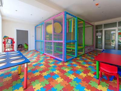 Area giochi interna luminosa con pavimento in puzzle di schiuma colorata, struttura gioco e piccoli tavoli.