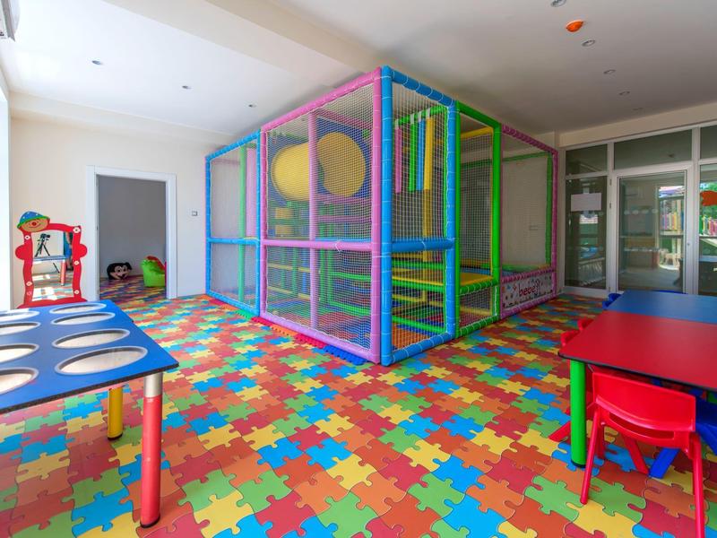 Area giochi interna luminosa con pavimento in puzzle di schiuma colorata, struttura gioco e piccoli tavoli.