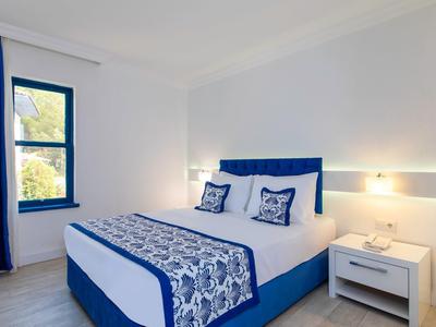 Moderní hotelový pokoj s modrou postelí, bílými stěnami a oknem se závěsy.