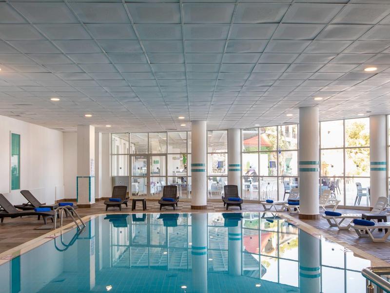 Vnitřní hotelový bazén s lehátky a velkými okny.