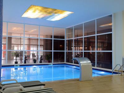Indoor swimming pool with glass walls, lounge chairs, and modern water feature under skylight.