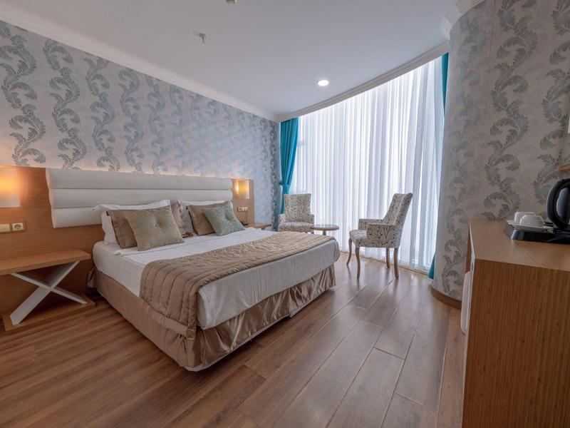 Hotel room with double bed, patterned wallpaper, wooden floor, and bright window with curtains.
