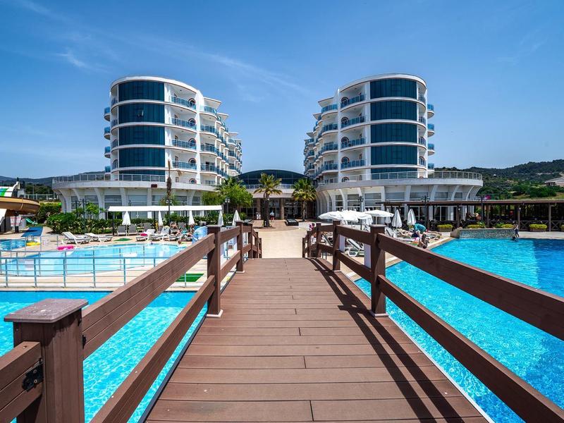 Wooden bridge leads to modern hotel buildings between large blue swimming pools under clear sky.