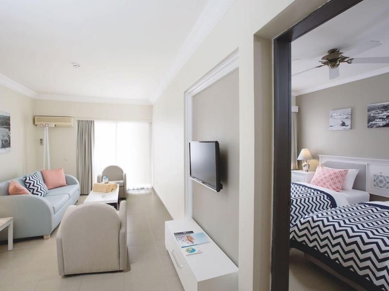 Appartement lumineux avec salon, canapé, TV et chambre adjacente avec lit double.