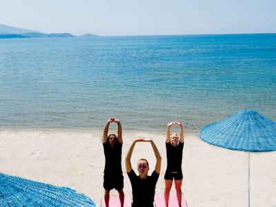 Drei Frauen in schwarzer Kleidung machen Yoga am Strand mit blauen Sonnenschirmen und Meer im Hintergrund.