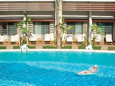 Ein Mann entspannt auf einer aufblasbaren Matte in einem hellblauen Pool vor einem Haus mit Liegestühlen.