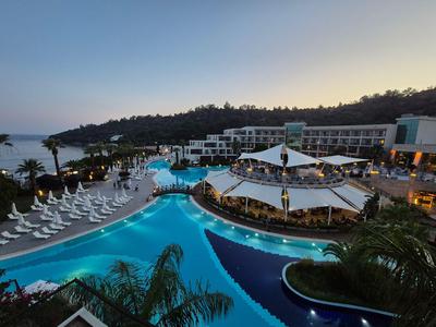 Großes Hotelresort mit beleuchteten Pools bei Dämmerung und weißen Liegestühlen am Meer.