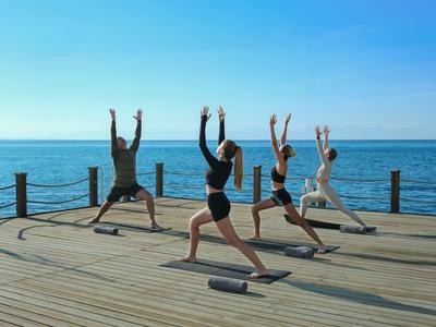 Vier Personen praktizieren Yoga auf einem Holzsteg mit Meerblick bei klarem Himmel.