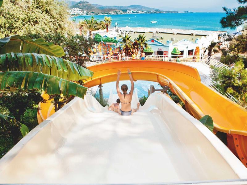 Person rutscht die weiße Wasserrutsche hinunter, umgeben von grüner Tropenvegetation und blauem Meer.
