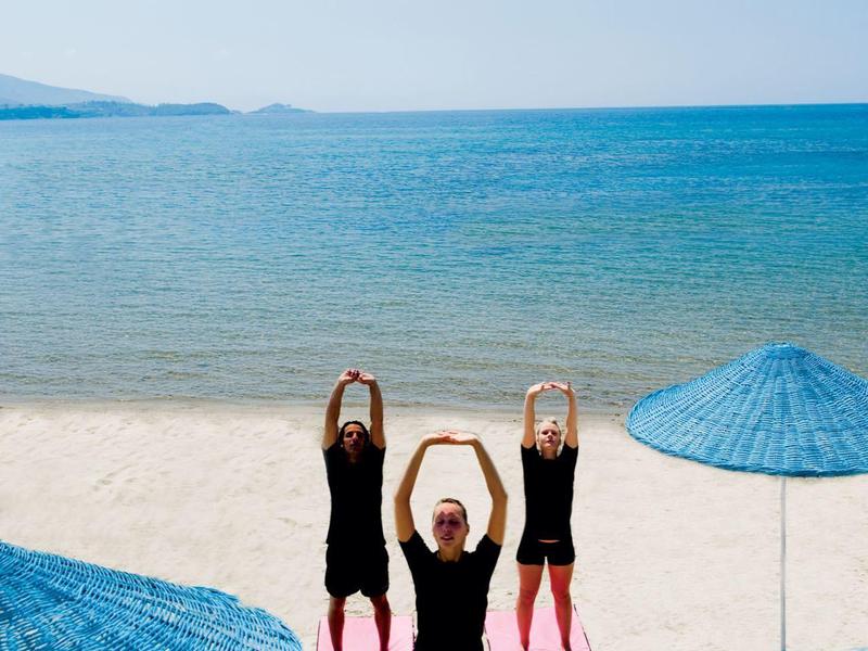 Drei Frauen in schwarzer Kleidung machen Yoga am Strand mit blauen Sonnenschirmen und Meer im Hintergrund.