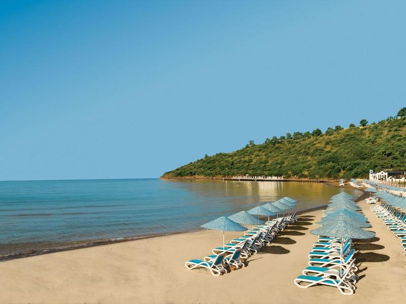 Leerer Sandstrand mit Reihen von blauen Liegestühlen und Sonnenschirmen, dahinter Hügel und blauer Himmel.