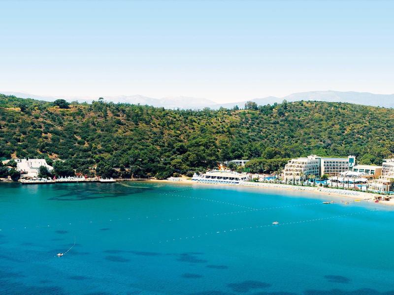 Klarer blauer Strand mit grün bewaldetem Hügel und mehreren Hotels am Ufer.