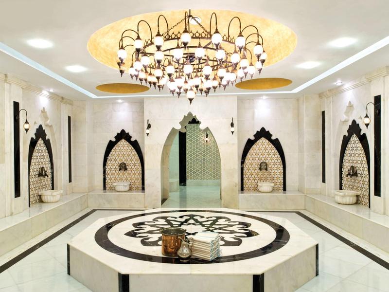 Ein heller, eleganter türkischer Hamam mit Marmor, verzierten Nischen und großem Kronleuchter.