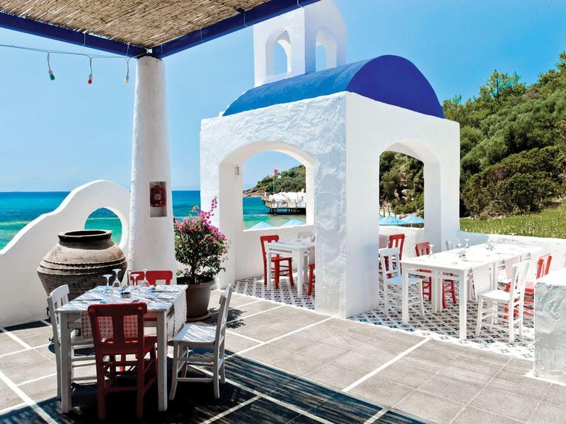 Offene Terrasse mit weiß-blauem Pavillon, weißen Stühlen, Holztischen und Meerblick unter blauem Himmel.