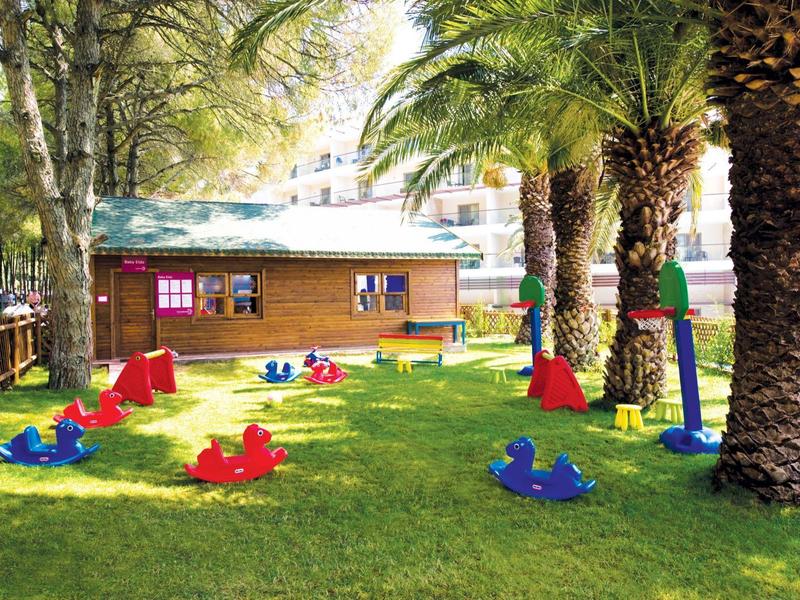 Grüner Spielplatz mit bunten Schaukeln, Palmen und einem Holzhäuschen im Hintergrund.