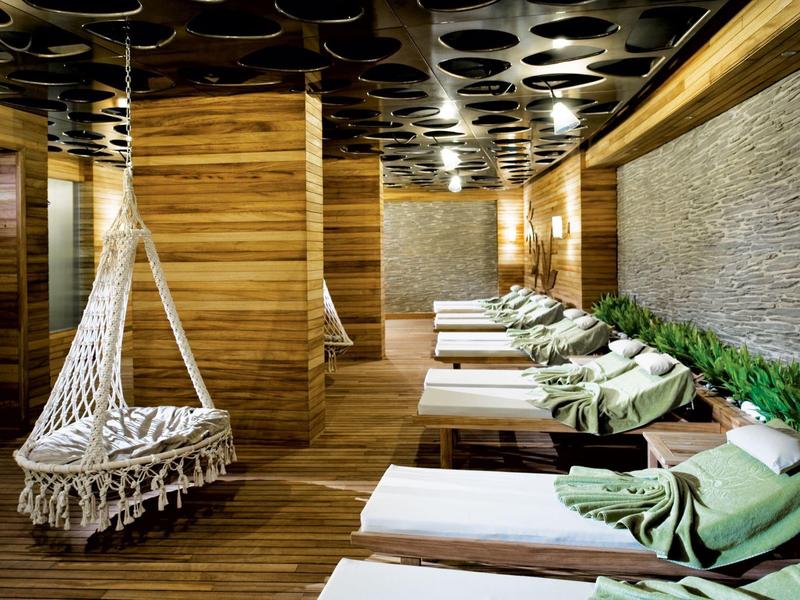 Entspannungsraum mit Holzdecke, Hängestuhl und Liegen mit grünen Handtüchern und Pflanzenwand.