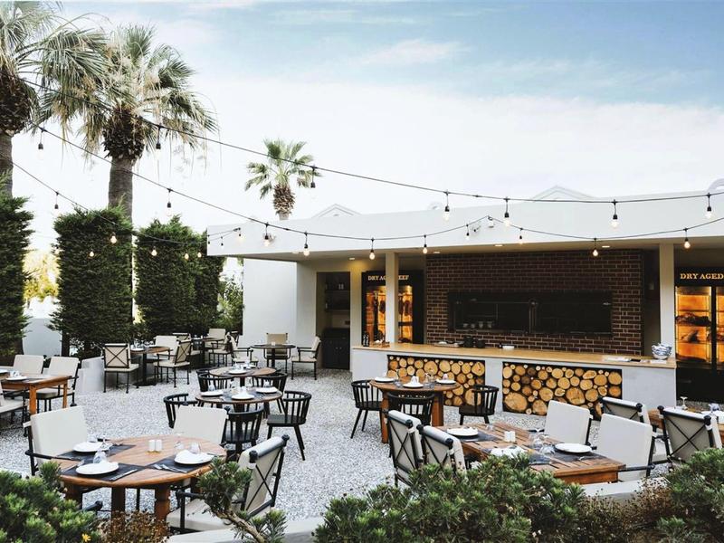 Moderner Außenbereich mit Tischen, Stühlen, Palmen und Lichterketten unter blauem Himmel.