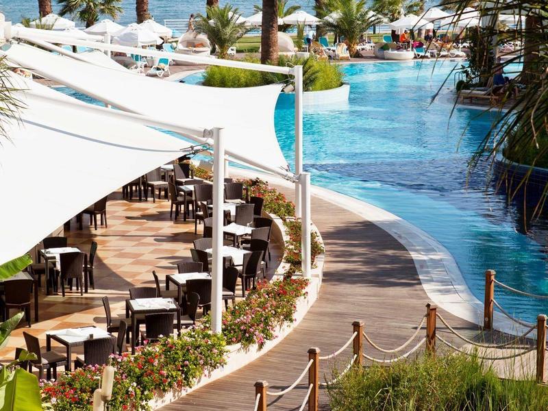Ein Pool mit blauem Wasser, umgeben von Palmen, Sitzbereichen und Blick aufs Meer im Hintergrund.
