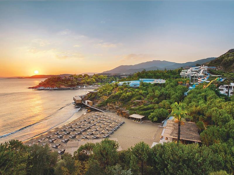 Resort costiero con ombrelloni, colline verdi e tramonto sul mare.