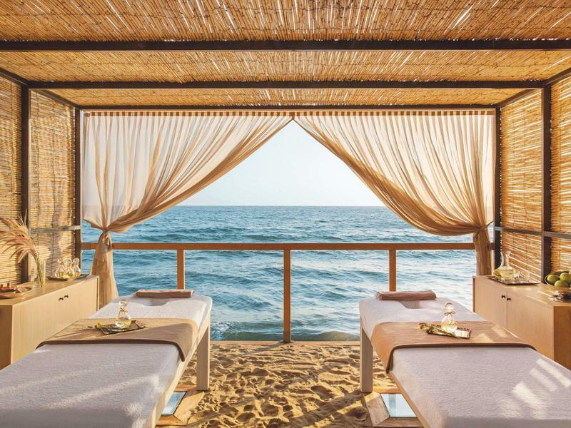 Area massaggi sul mare con due lettini sotto un tetto di paglia e tende leggere.