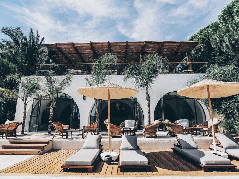 Area piscina di hotel moderno con lettini, ombrelloni e palme sotto un cielo parzialmente nuvoloso.