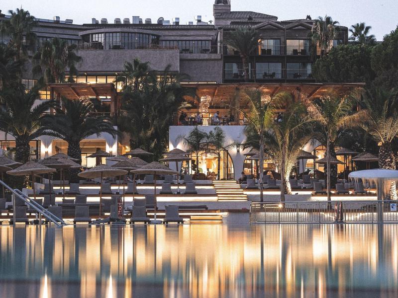 Hotel di lusso al crepuscolo con lettini illuminati a bordo piscina e palme riflesse nell'acqua.