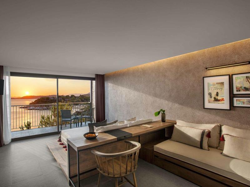 Camera d'hotel moderna con divano, scrivania e grande finestra con vista al tramonto sull'oceano.