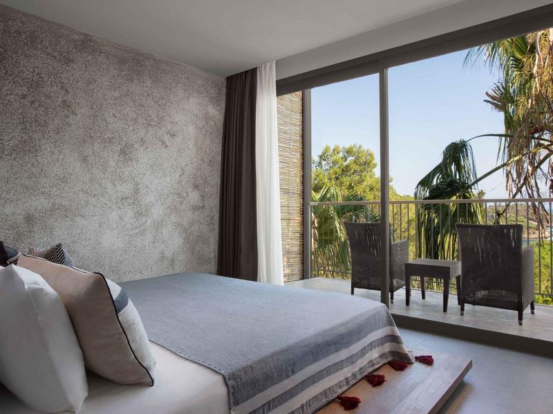 Camera d'hotel moderna con letto grande e balcone con vista su vegetazione e palme.