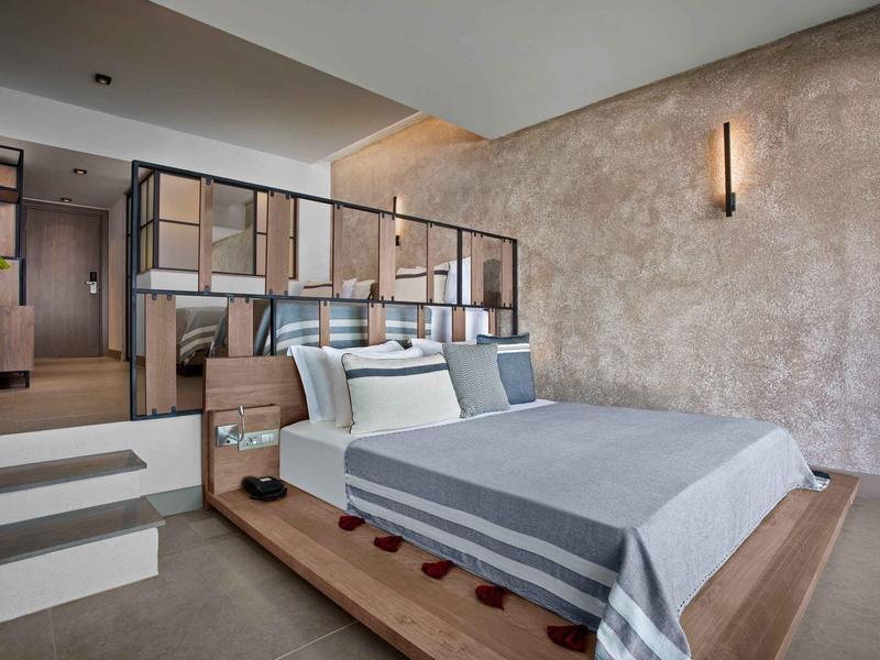 Camera d'albergo moderna con letto grigio, piattaforma in legno e parete beige strutturata con lampada da parete.