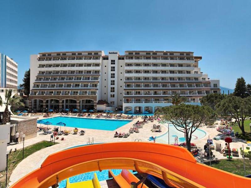 Hotel con grandi piscine, scivoli d'acqua e terrazza solarium sotto un cielo blu chiaro.