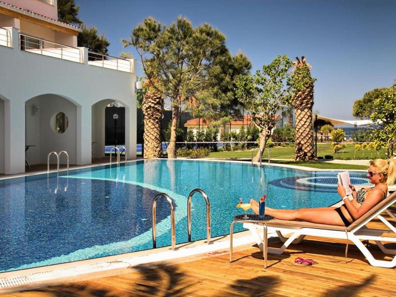 Area piscina di un hotel con terrazza solarium, lettini e palme sotto un cielo limpido.