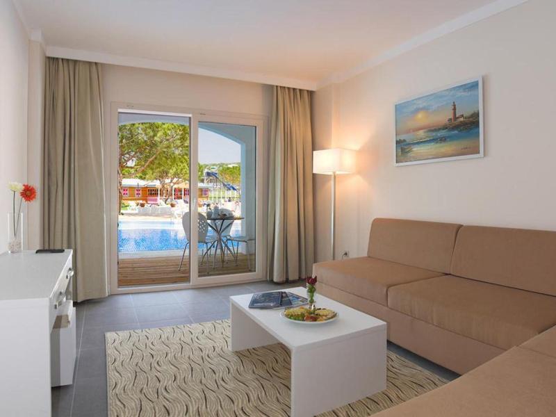 Camera d'hotel luminosa con divano, tavolino e vista sulla piscina attraverso grandi porte in vetro.