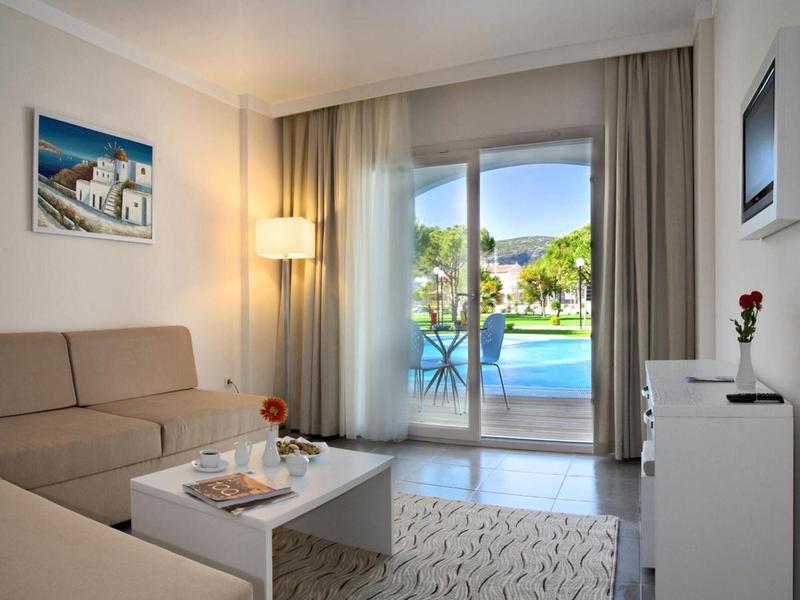 Camera d'hotel luminosa con divano, tavolino e vista sulla piscina attraverso grandi porte scorrevoli in vetro.