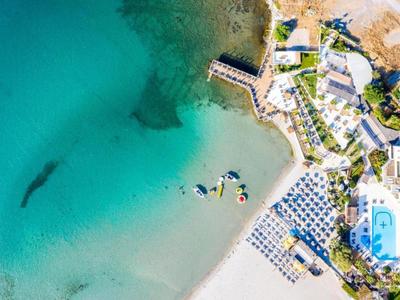 Luftaufnahme eines Strandes mit türkisfarbenem Wasser, Sonnenliegen, Pool und angrenzenden Gebäuden.