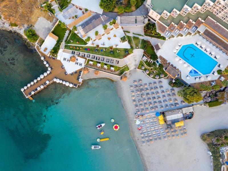 Vista aérea de un hotel en la playa con piscina, tumbonas y mar azul claro con equipos de deportes acuáticos.