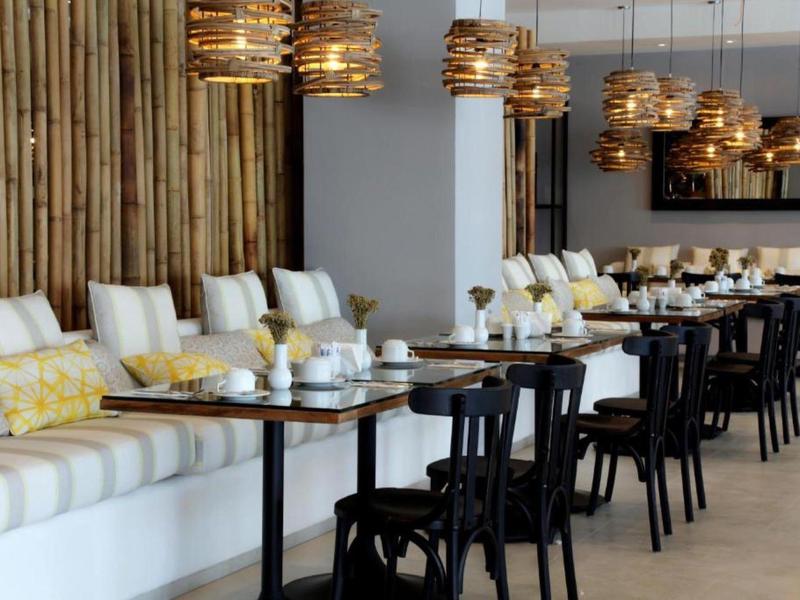 Área de comedor moderna con mesas de madera, sillas negras y lámparas colgantes en espacio claro.