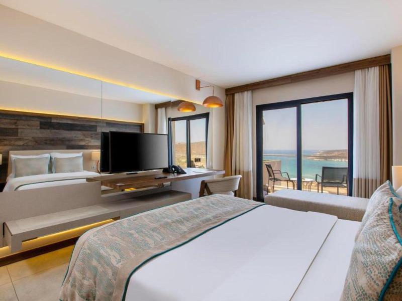 Habitación de hotel moderna con cama doble, televisión y vista al mar a través de grandes ventanales.