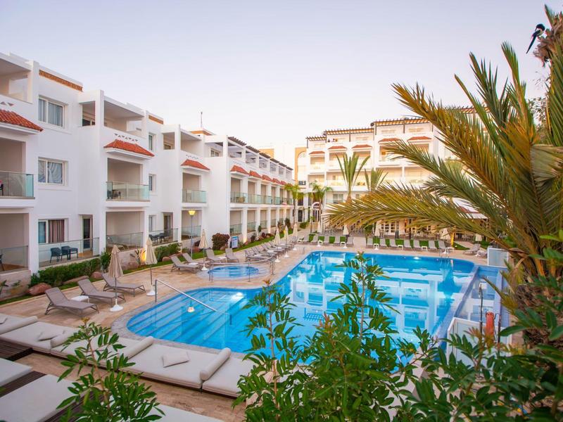 Moderner Außenpool mit Liegestühlen, umgeben von weißen mehrstöckigen Hotelgebäuden und Palmen.